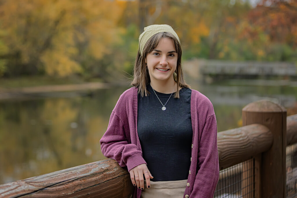 Annaliese Dorchinecz is a young white woman. In this picture, she is leaning against the rail of a wooden bridge and is beside a lake and trees of many colors during the autumn season. She is wearing a faded red cardigan, a black shirt, a green bandana, and khaki pants.