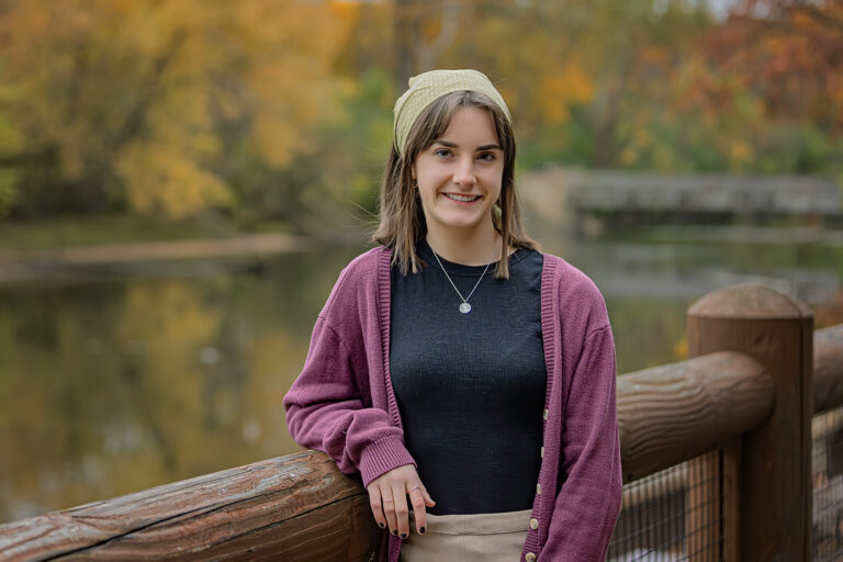 Annaliese Dorchinecz is a young white woman. In this picture, she is leaning against the rail of a wooden bridge and is beside a lake and trees of many colors during the autumn season. She is wearing a faded red cardigan, a black shirt, a green bandana, and khaki pants.